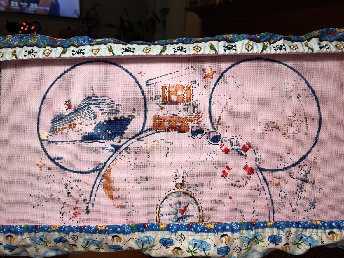 Anne Conrad&rsquo;s &ldquo;Magical Cruise&rdquo; cross stitch in progress on pale pink fabric in a scroll frame — the cruise ship stitched in the left ear, suitcases stacked at the top, and the compass rose taking shape below