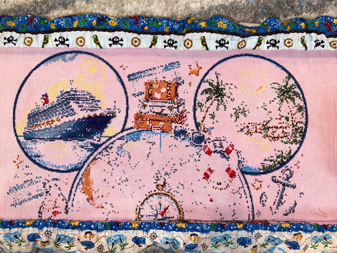 Anne Conrad’s “Magical Cruise” cross stitch in progress on pink hand-dyed fabric in a scroll frame with pirate-print guards — the cruise ship stitched in the left ear, the stacked luggage between the ears, the island and palm trees in the right ear, and the compass rose and anchor taking shape below
