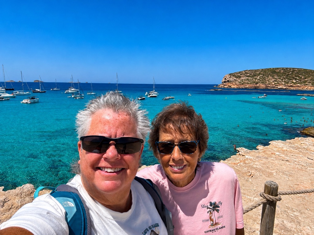 Anne Conrad and her wife Kathy in Ibiza, with sailboats anchored in the turquoise bay behind them