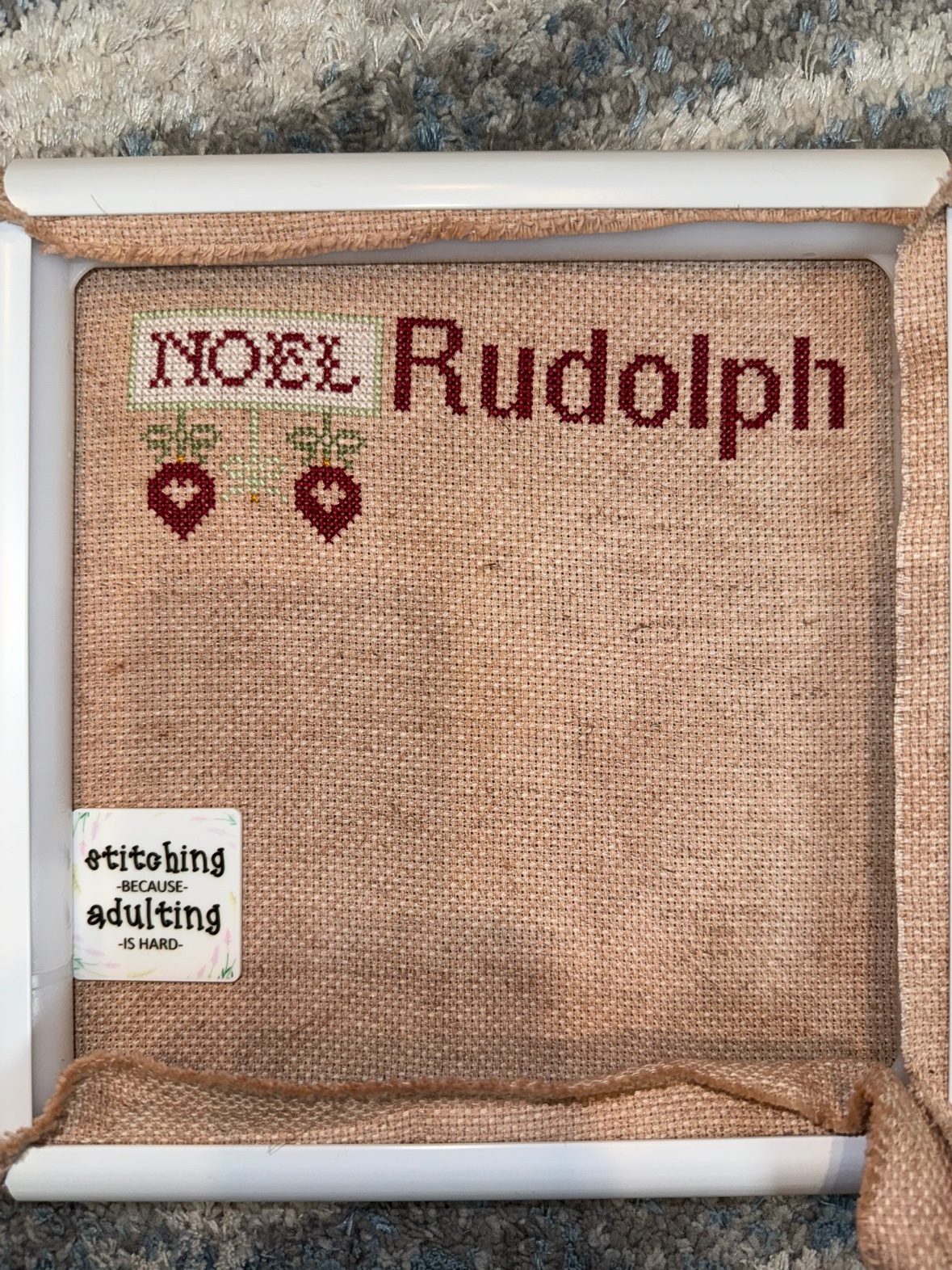 Kathy’s new start, “Christmas Magic” by Primrose Cottage Stitches, on warm tan hand-dyed fabric in a white frame — NOEL stitched with two red heart ornaments hanging below it and “Rudolph” started in red beside it, with a needle minder reading “stitching because adulting is hard”