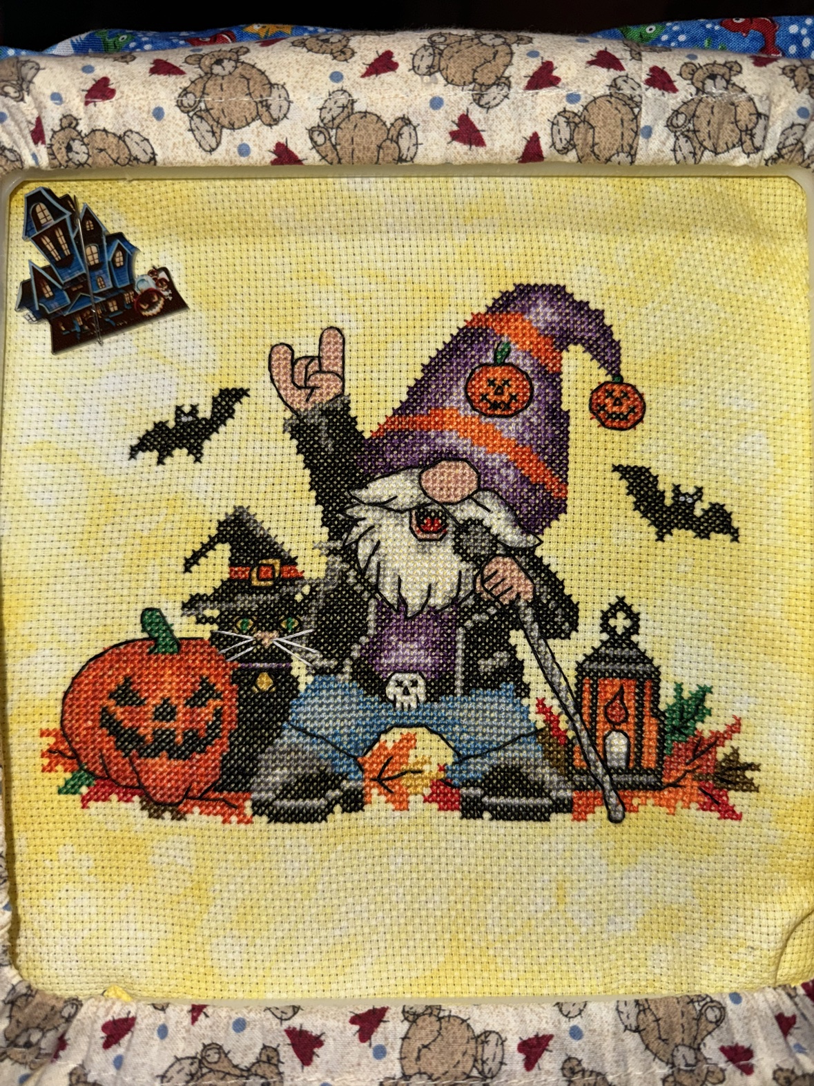 Kathy&rsquo;s finished cross-stitch, &ldquo;Totally Rocking Halloween&rdquo; by Michaela Learner — a rock-on gnome with a microphone stand, a black cat in a witch hat, a jack-o&rsquo;-lantern and a lantern on yellow mottled fabric, the haunted-house needle minder still in the corner